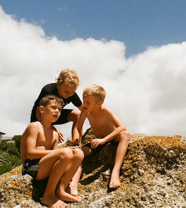 Color photograph of three young boys wearing bathing suits sitting on a large rock.
