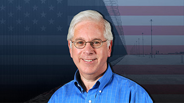Andrew I. Schoenholtz, a middle-aged white man in a blue shirt, smiles in front of a digital background of the American flag overlayed on top of a picture from the southern border with barbed wire fencing and communications towers.